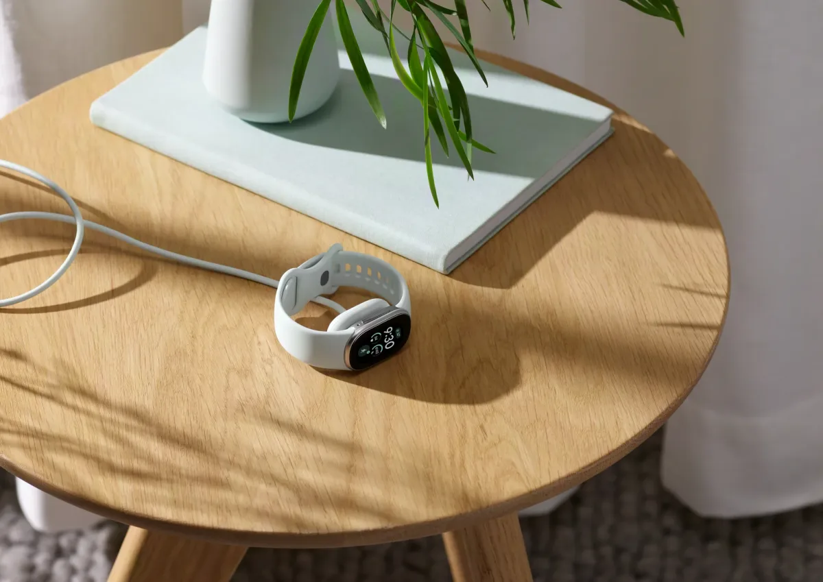 Fitbit Edge Fitness-Tracker mit weißem Armband liegt beim Laden auf einem Holztisch